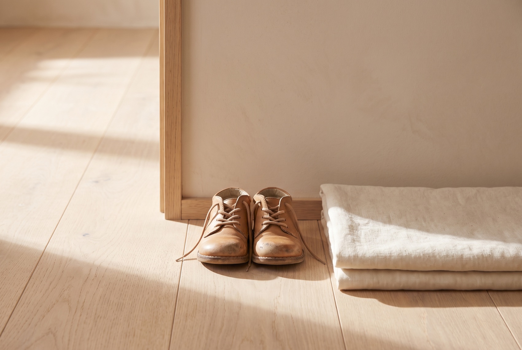 A pair of small worn leather shoes placed neatly side by side on a sunlit wooden floor by a door.